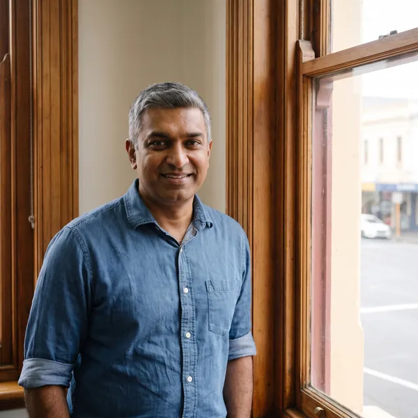 Rajiv Menon, in a blue denim shirt, standing by the timber-framed front window of the office, smiling.