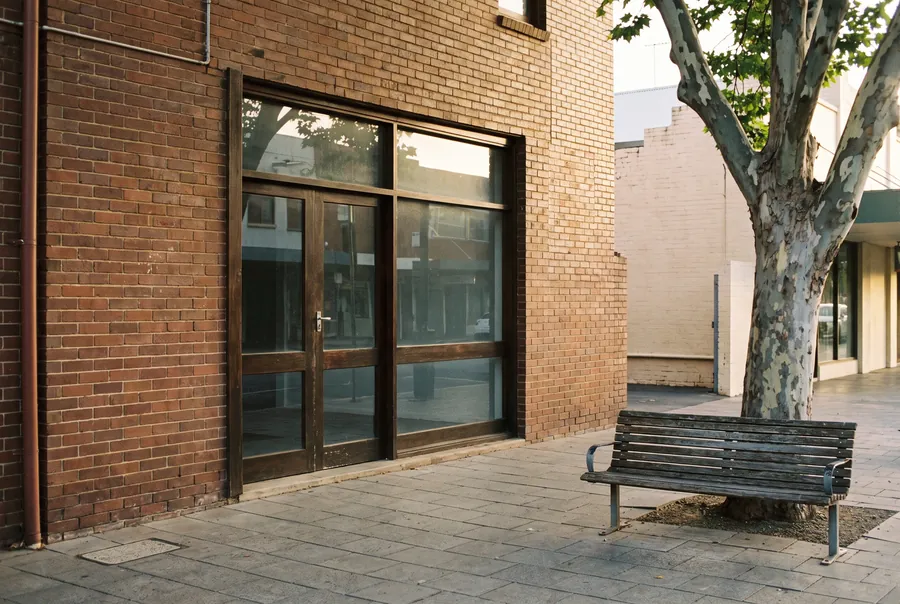 The office from the footpath: a red-brick 1920s shopfront with tall timber-framed windows and a glass door, a bench under a plane tree beside it.