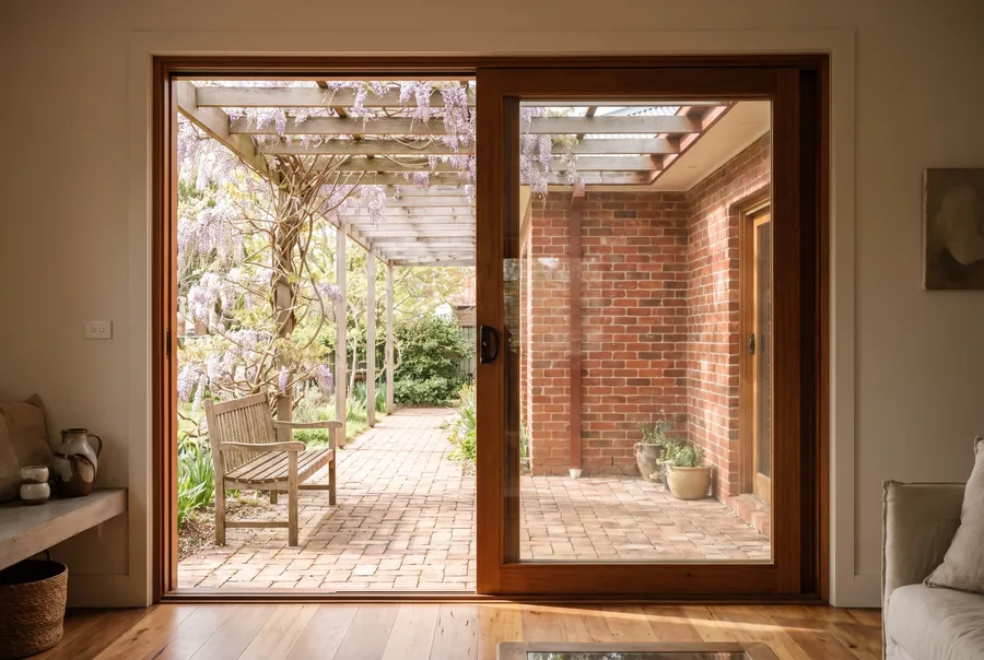 Looking out through a wide timber sliding door to a brick-paved courtyard under a pergola covered in wisteria.