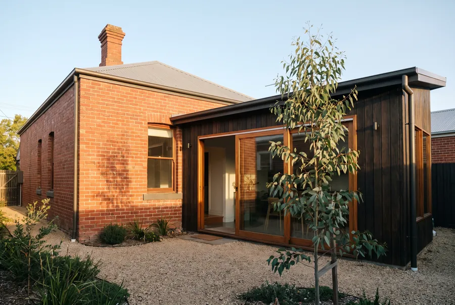 A low, dark-stained timber addition with timber-framed glass doors, set back from the corner of an old red-brick cottage.