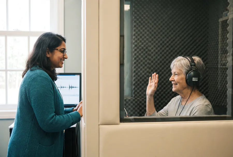 Through the sound booth window, a woman in headphones raises her hand to show she heard a tone, and the clinician outside smiles back.
