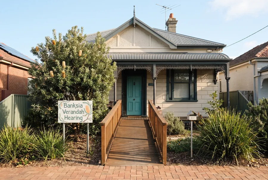 A Federation cottage with a teal front door, a timber ramp up to the verandah, and the Banksia Verandah Hearing sign beside a flowering banksia.