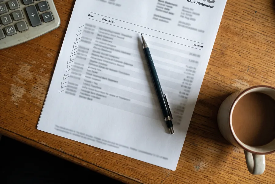 A printed bank statement on a wooden desk, ticked down the margin in pencil, with a pencil lying across it, a calculator and a mug of coffee.