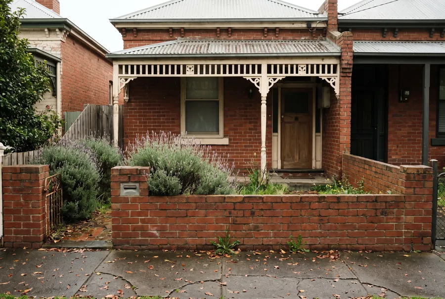 A single-fronted red-brick cottage with a white iron-lace verandah, a timber front door and lavender behind a low brick fence.