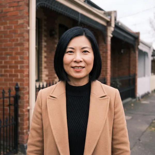 Vivian Liang, smiling, in a camel coat over a black jumper, outside a row of brick terraces with iron fences.