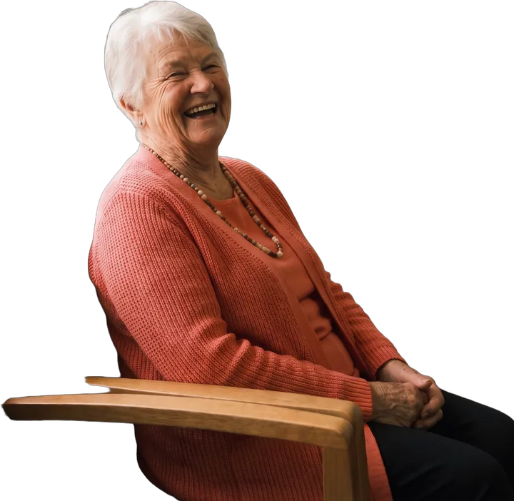 Maureen, a woman in her seventies with short white hair and a coral cardigan, laughing in a timber chair.