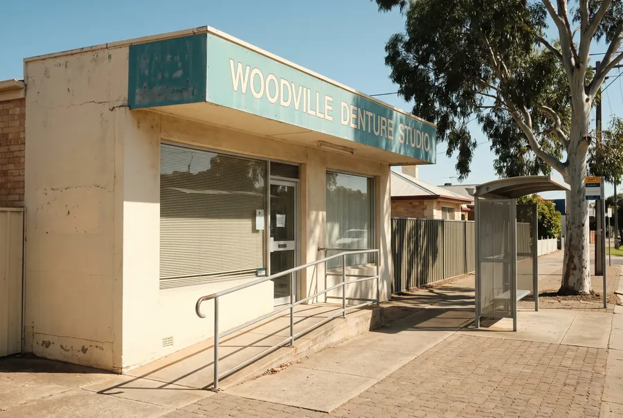 The studio on Port Road: a cream building with a teal Woodville Denture Studio sign, a ramp with handrails to the door, and a bus shelter out front.