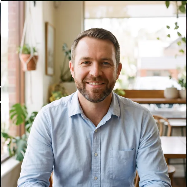 Ben Carter, smiling, in a pressed light blue button-up shirt at a cafe table, with indoor plants behind him.