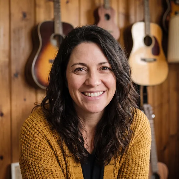 Gina Marino, smiling, in a mustard cardigan in front of guitars on a timber wall.