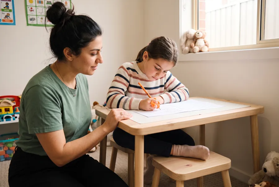 Leila sits beside a girl writing at a small wooden table. The girl’s pencil has an orange grip and her feet rest on a stool.