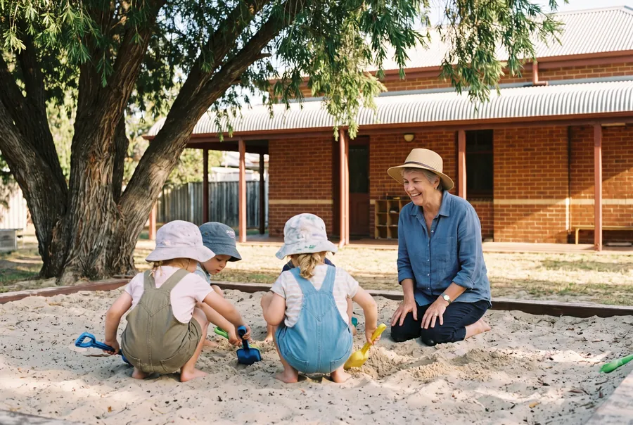 Glenys Pike, the education assistant, kneeling in the sandpit and laughing with three children in sun hats as they dig.