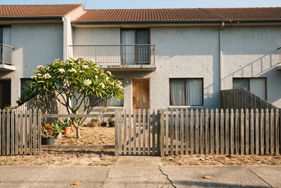 Two-storey rendered townhouse behind a timber picket fence, with a frangipani and potted plants in the front garden.