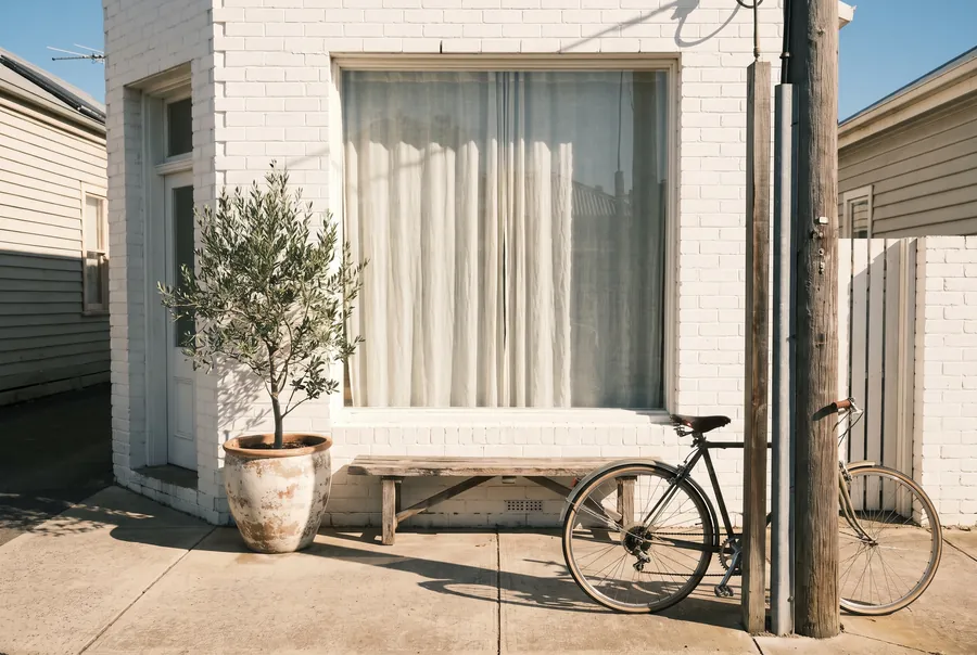 A white-brick shopfront with a big curtained window, a door on the left with one step up, a potted olive tree and a bench out front.