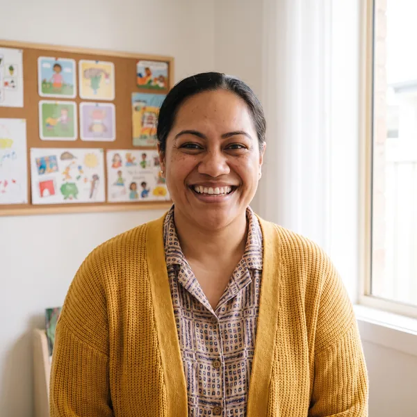 Mele Fifita smiling in a yellow cardigan, picture cards pinned on the wall behind her.