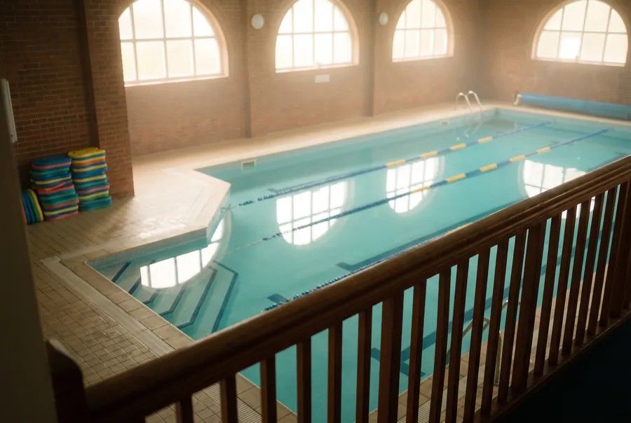 The indoor teaching pool seen from the viewing balcony: three lanes, wide steps in the shallow corner and stacks of kickboards on the deck.